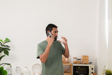 Happy young Asian man using a smartphone drinking clean water in the morning immediately after waking up. Healthy smart man cleanses his body by drinking water after waking up first.