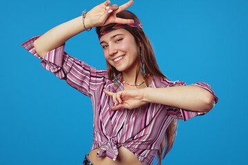 Obraz na płótnie Canvas Positive hippie woman with headband makes peace gesture with both hands smiling happy dressed in pink striped shirt, isolated over blue background.