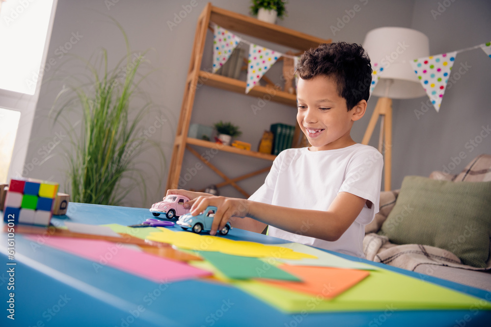 Wall mural photo of cheerful positive little boy dressed white t-shirt sitting table enjoying game indoors hous