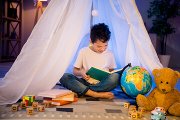 photo of positive clever geek nerd boy sit in homemade tent read interesting book in evening playroom indoors