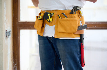 Cropped view of the man in white T-shirt and blue jeans with yellow tool belt near concrete or cement wall and wide light window.