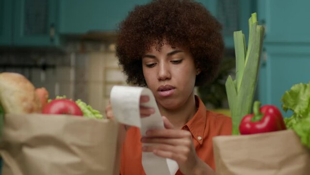 20s Woman Checking Grocery Receipt. Black female looking at grocery bags full of fresh food.