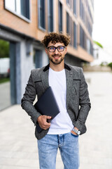 Handsome young businessman holding laptop while using his phone and standing on the street