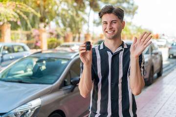 Young handsome man holding car keys at outdoors saluting with hand with happy expression