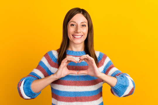 Image of attractive young woman with long brown hair smiling and showing heart shape with fingers isolated over yellow background