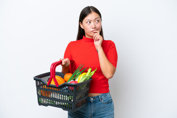 Fototapeta na wymiar Young Asian woman holding a shopping basket full of food isolated on white background having doubts and thinking
