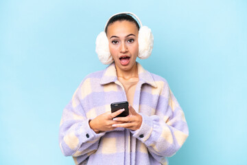 Young Arab woman wearing winter muffs isolated on blue background surprised and sending a message