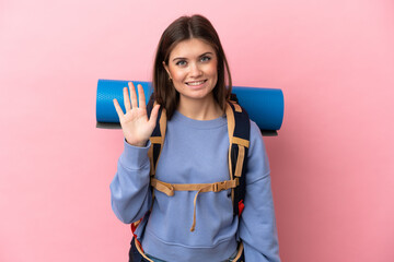Young mountaineer woman with a big backpack isolated on pink background saluting with hand with happy expression