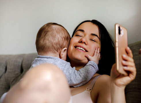 mother and baby son boy hugging kissing caresses cuddling sitting on sofa talking at smartphone.happy woman in maternity with infant toddler child.portrait,smiling female adorable new born pregnant 