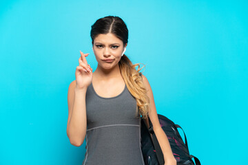 Young sport Russian girl with sport bag isolated on blue background with fingers crossing and wishing the best