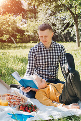 Family picnic. Dad and son in a green garden in sunny weather on a picnic eating pizza and reading book. Front view
