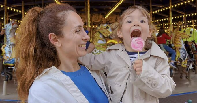 A little girl in her mother's arms eats a colorful candy in an amusement park. A three-year-old child licks a candy at a party. Family portrait mom with cute daughter