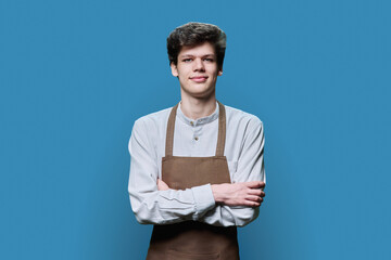 Fototapeta premium Portrait of young guy in apron with crossed arms on blue background