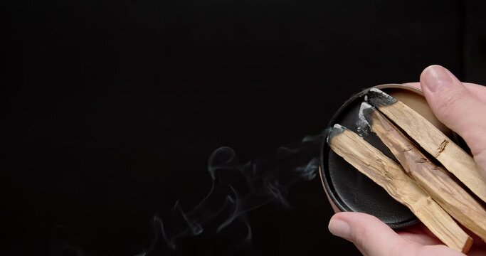 Burning stick of palo santo in hands on black background. Person holding Palo Santo in hands, spreading smoke. High quality video clip. Close up 4k footage