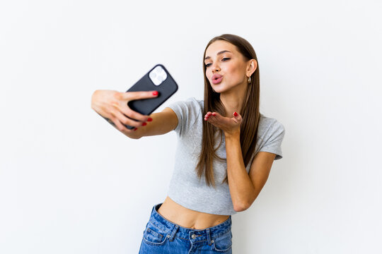 Happy flirting young woman taking pictures of herself at smart phone blowing a kiss, over white background