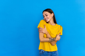 Young woman standing over blue background hugging oneself happy and positive, smiling confident.