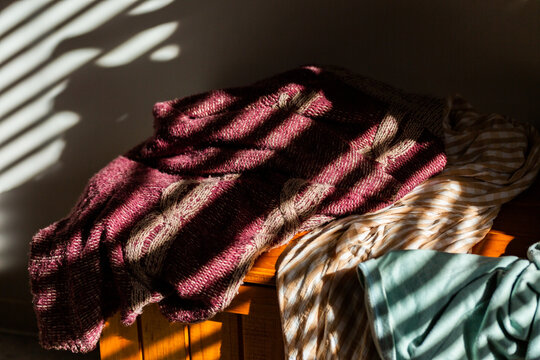 Light and shadow in bars falling on pastel coloured warm winter clothing draped over blanket box