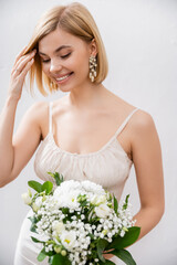 cheerful and blonde bride in wedding dress holding bouquet on grey background, white flowers, bridal accessories, happiness, special occasion,   beautiful, feminine, blissful