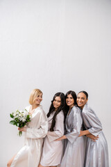 positivity, happy bride holding bouquet of flowers and standing near cheerful interracial bridesmaids on grey background, racial diversity, silk robes, fashion, friendship, brunette and blonde women - obrazy, fototapety, plakaty