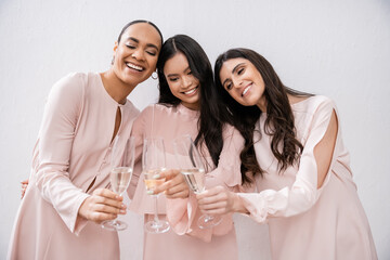three multicultural bridesmaids, pretty women in pastel pink dresses clinking glasses of champagne on grey background, cultural diversity, fashion, celebration, cheers