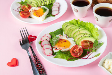Healthy romantic breakfast - two portions heart-shaped fried eggs with avocado, spinach, cherry tomatoes and radish on plates on pink background