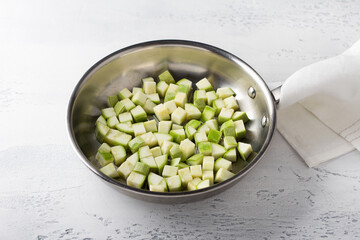 Raw zucchini diced in a frying pan with oil on a light gray table. Cooking delicious vegan food