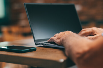 Defocused senior man sitting at bar table using laptop and mobile phone. Mature caucasian man typing on keyboard in business work