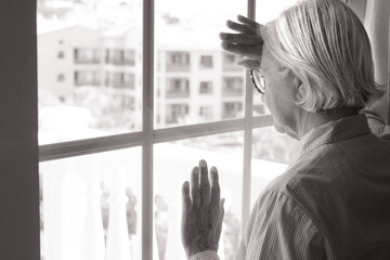 Black and white portrait of sad melancholy senior caucasian woman at home looking out the window,...
