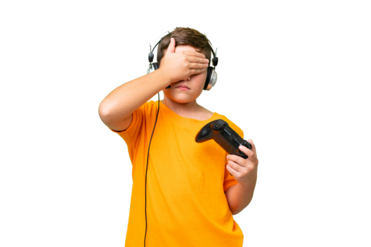 Little caucasian kid playing with a video game controller over isolated chroma key background covering eyes by hands. Do not want to see something