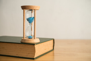 Hourglass and textbook on wooden table in classroom with white wall background copy space vintage style. Time to study, back to school, education and self development concept.