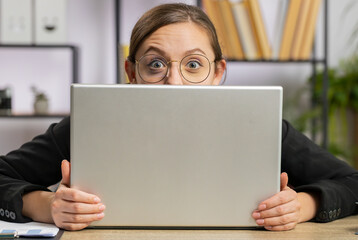 Confident business woman hiding behind laptop computer, looking at camera, spying his colleagues working, peeping. Professional freelancer girl looking from behind computer with cunning eyes glance - obrazy, fototapety, plakaty