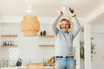 Ecstatic excited grey-haired man in casual and wireless headphones enjoy spare time, listen favorite music and dancing in front kitchen room at home. Elderly weekend activities lifestyle concept
