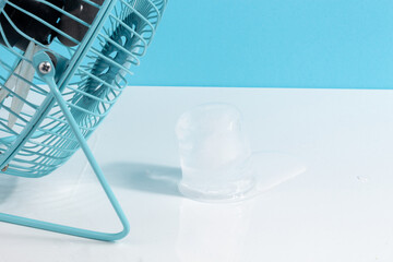 High temperature concept with a little blue fan cooling an ice cube melting on white and blue...