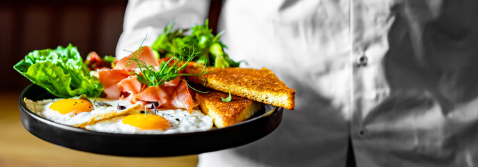 chef hold plate with fried eggs, ham, salad and hummus in cafe