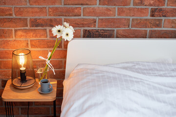 Reading lamps near the bed in the bedroom. Concept of interior design in modern loft apartment. Cozy bedroom with home decor, bedside nightstand with shining lamp, bouquet of flowers and coffee cup.