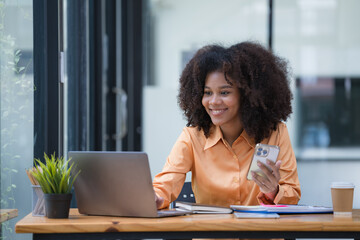 Portrait of a beautiful confident businesswoman using a laptop computer holding a mobile phone sitting in a modern office. Smiling African American freelancer working online from home.  - obrazy, fototapety, plakaty