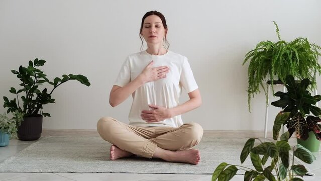 Pretty female sitting on the floor in lotus posture with hands on her chest and belly doing pranayama techniques. Breathing techniques. Doing yoga at home. Self care and positive emotions.