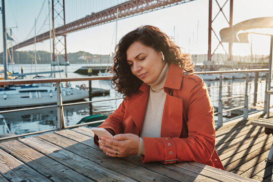  Pregnant brunette woman in an orange coat sitting on the waterfront at a table with a phone in hand at sunset in front of the pier with yachts and the bridge in Lisbon