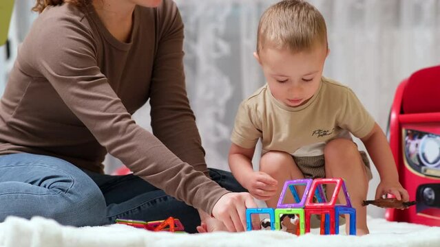 The nanny spends time in a modern cozy house with a little boy. Loving caring mother plays with her preschool son, builds a tower of magnetic blocks. Child development, play time concept
