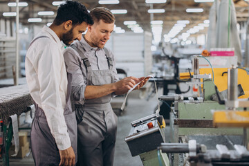 A male inspector or operator of a workshop for the production of aluminum and plastic wreaths trains an intern. International team of men working together near a machine in a factory.