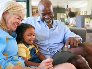 Grandparents And Granddaughter With Digital Tablet Sitting On Sofa At Home Together