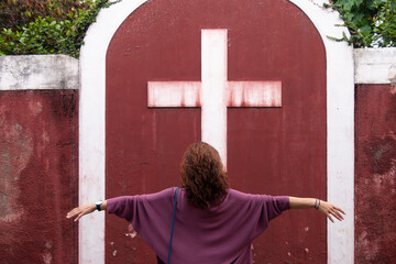 Una mujer de espalada y frente a una cruz con los brazos abiertos.