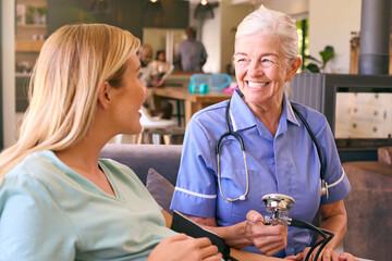 Senior Midwife Visiting Pregnant Woman At Home Taking Blood Pressure With Family In Background