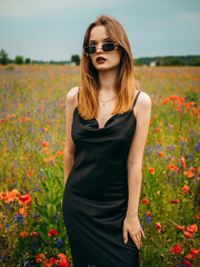 Beautiful young girl in a black evening dress and sunglasses posing against a poppy field on a cloudy summer day. Portrait of a female model outdoors. Rainy weather. Gray clouds. Vertical shot.