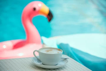 Fotobehang A cup of coffee by the pool and an inflatable pink flamingo in the water. © Ann Stryzhekin