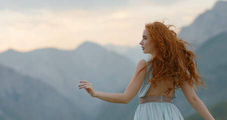 Fototapeta na wymiar Beautiful girl with red hair in white dress looking at landscape in mountains while wind blows her hair - freedom, inspiration cinematic. Copy space