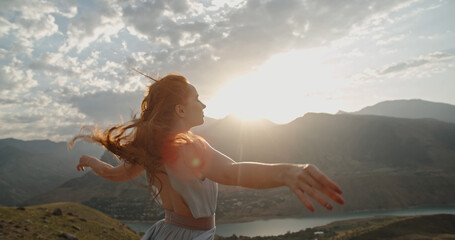Woman in white dress standing on top of a mountain with raised hands while wind is blowing her...