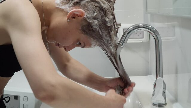 Caucasian girl washes her head with soap in the bathroom in the sink. European woman in a sports top bra washes her blond hair with shampoo with foam under a tap with water at home in an apartment