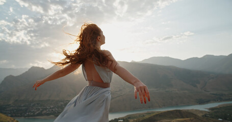 Woman in white dress standing on top of a mountain with raised hands while wind is blowing her...