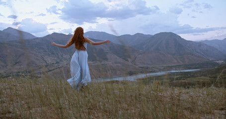 Young beautiful girl with red hair wearing white dress walking on top of a mountain facing wind blowing her hair and dress - freedom, adventure, harmony 